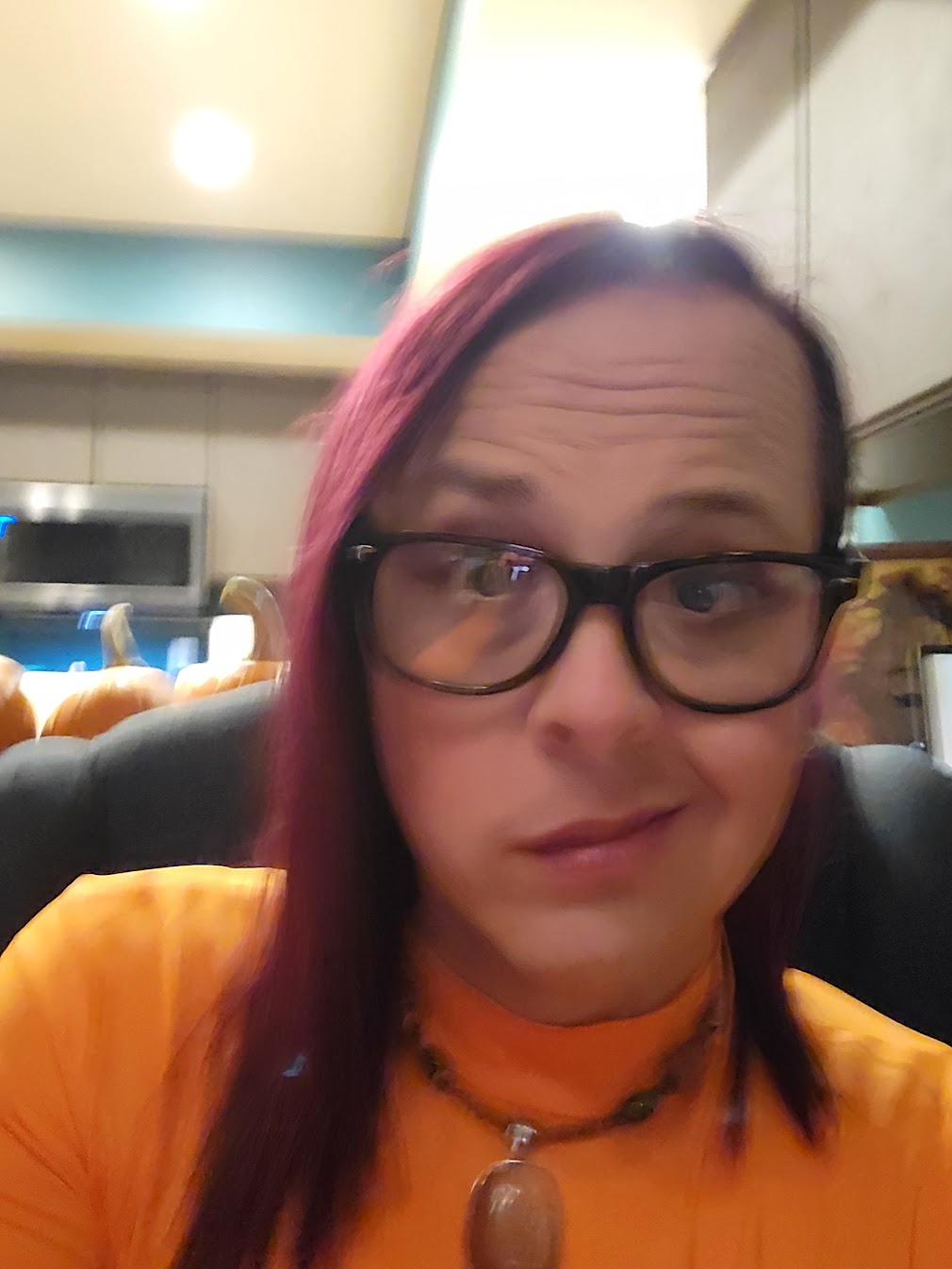 Zoe headshot - a woman with purple shoulder length hair swept to one side and a sideshave on the other, she is caucasian with blue eyes and a smirk and purple lipstick, sitting in front of an ornate headboard for a bed. 